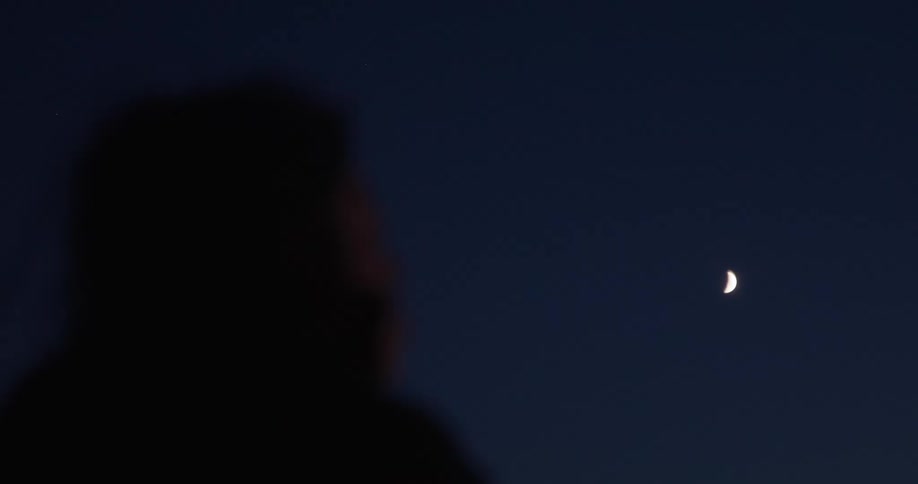 A person's dark silhouette is seen in the foreground looking up at a small crescent moon in the deep blue twilight sky