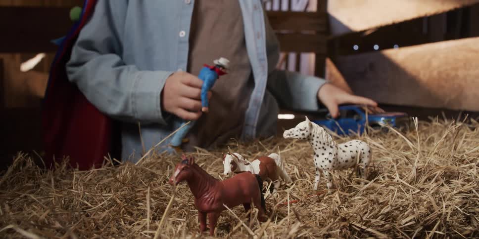 A young boy plays with his toy horses, cowboy, and truck in a pile of hay inside a rustic wooden barn
