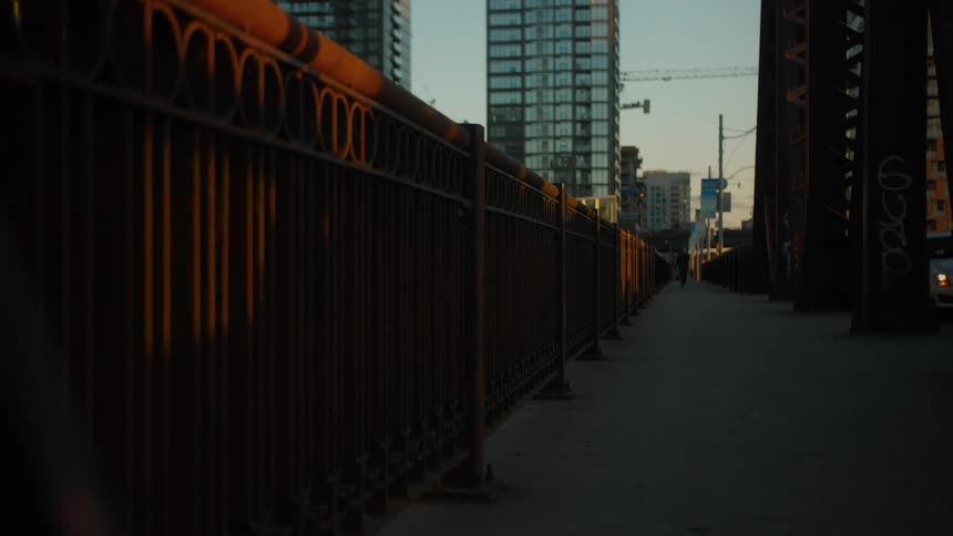 Golden hour sunlight casts moving shadows from passing cars along the railing of an urban bridge walkway