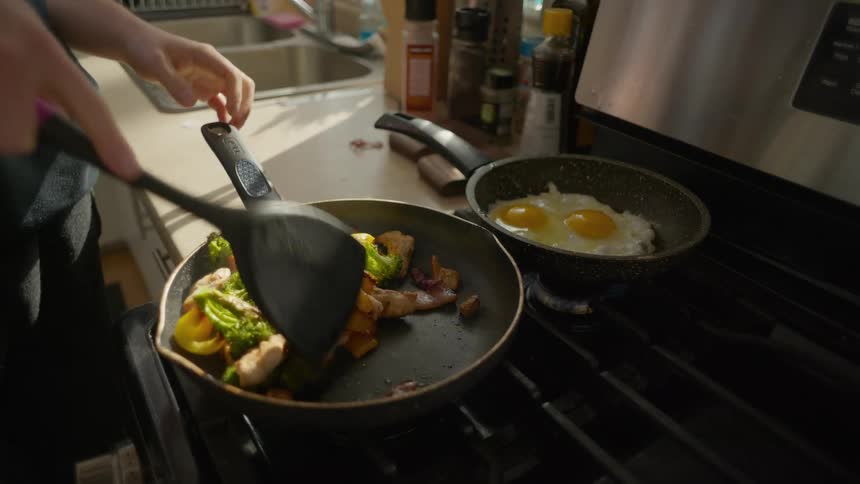 Slow motion close-up of a person stir-frying a healthy meal of chicken and vegetables while eggs cook on a sunlit stovetop