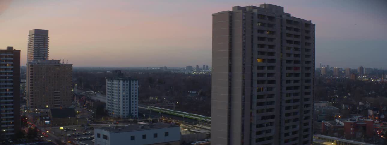 A green commuter train travels through a cityscape with apartment buildings during a peaceful twilight
