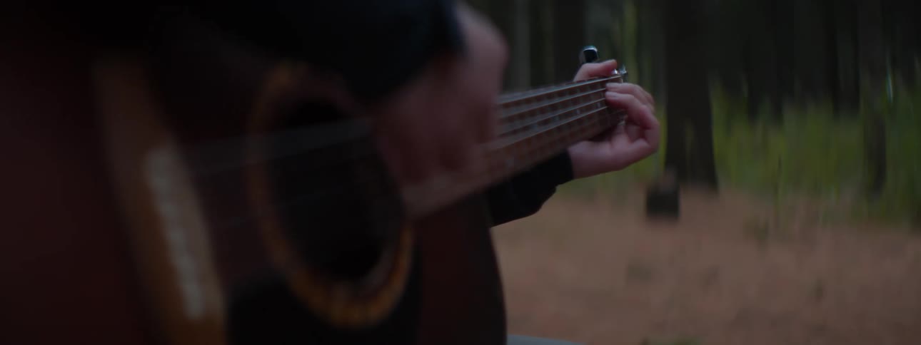 A close-up of a person's hands strumming an acoustic guitar while sitting in a tranquil forest