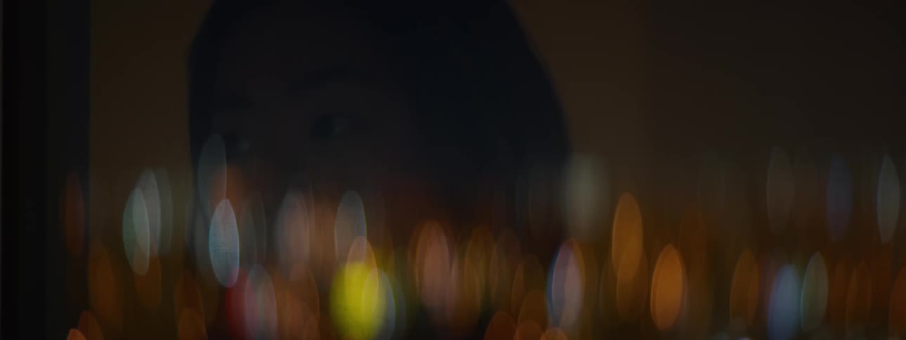 A person's contemplative reflection is faintly seen in a window against a beautiful backdrop of blurry city lights at night