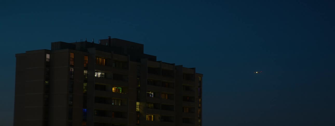 A long shot shows an airplane flying across the night sky behind a tall apartment building with its windows illuminated