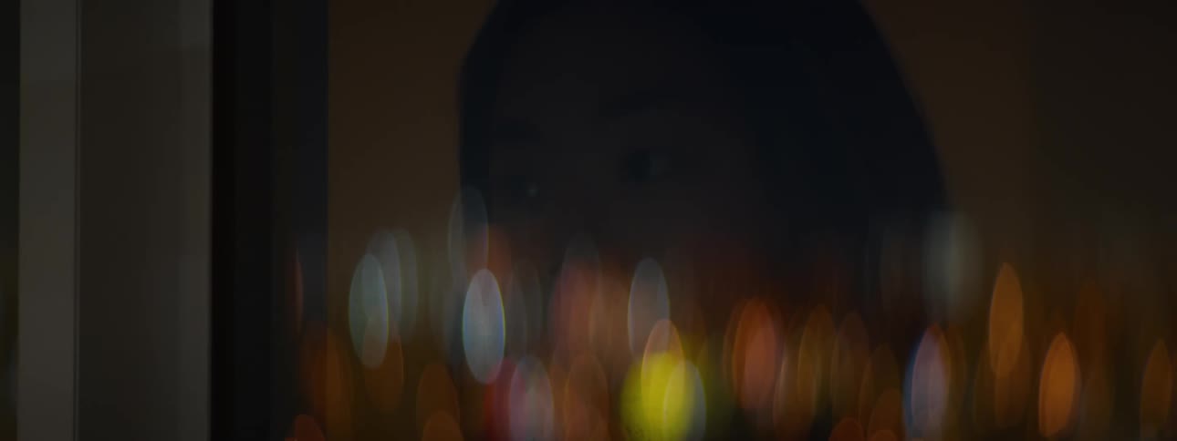 A contemplative person's eyes are reflected in a window with a vibrant bokeh of out-of-focus city lights at night
