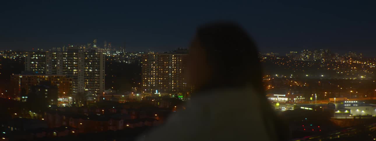 An out-of-focus person contemplates the glowing city lights and bustling highway traffic from a high vantage point at night