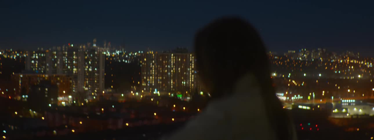 A woman looks out over the city skyline at night as the camera pulls focus from bokeh lights to the distant buildings