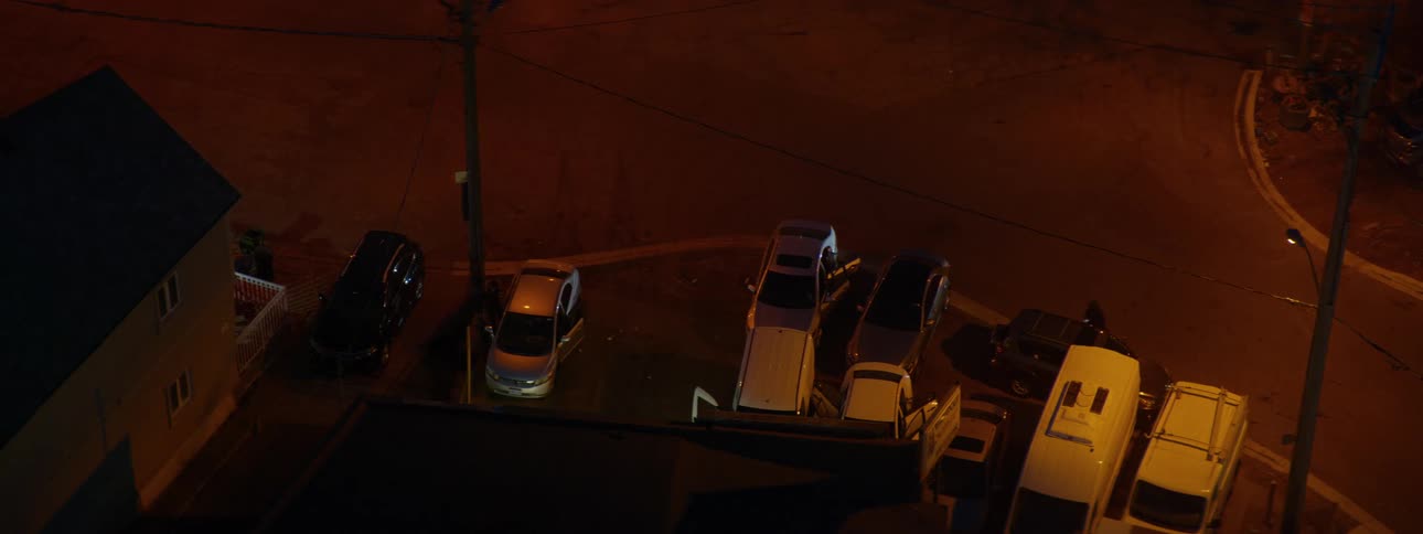 An overhead shot captures a quiet urban parking lot at night as a person walks to their car