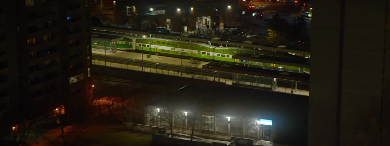 A high-angle view shows a green and white commuter train leaving the brightly lit station in the city at night