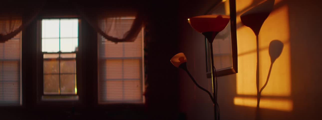 Golden hour sunlight streams into a dark room, illuminating a floor lamp and causing a sheer curtain to blow gently in the breeze