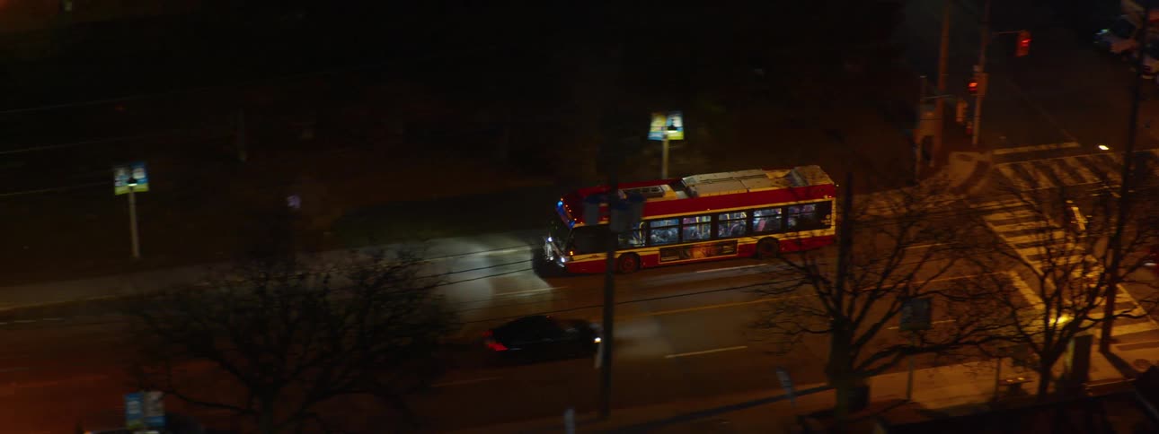 A red city bus drives through a lit urban intersection at night as seen from a high angle