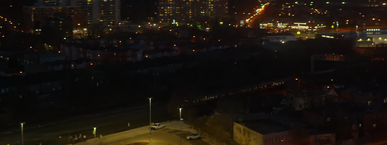 High angle view of a commuter train leaving a station at night, pulling back to reveal the wider suburban cityscape with glowing lights