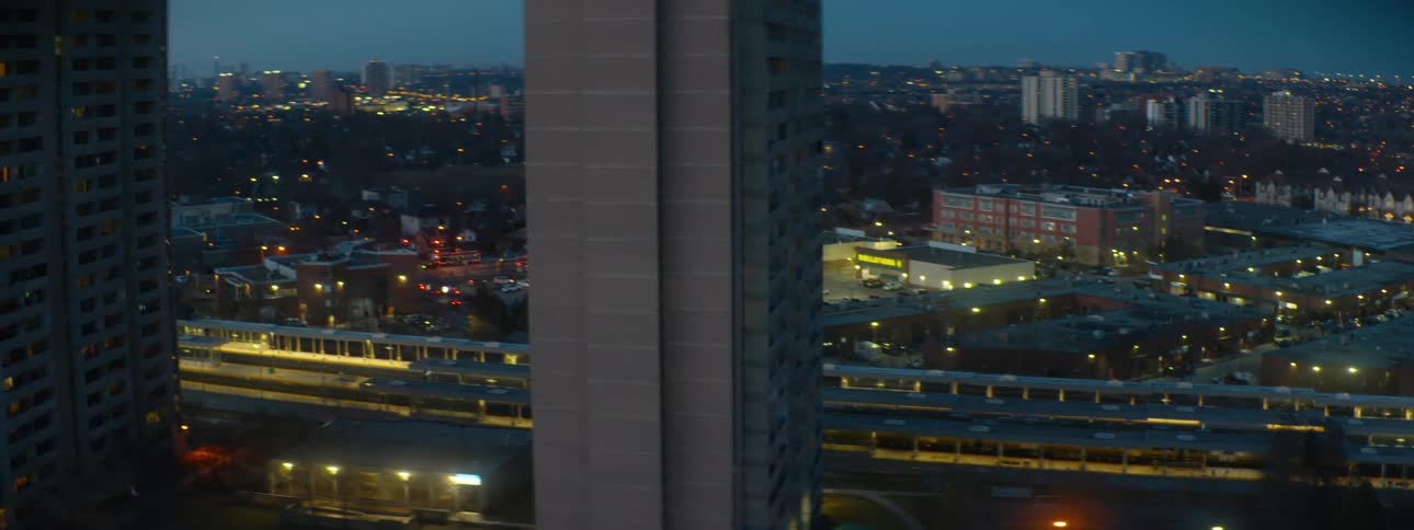 An evening view of a city transit station pans left and tilts up to reveal a sprawling urban landscape framed by two apartment buildings