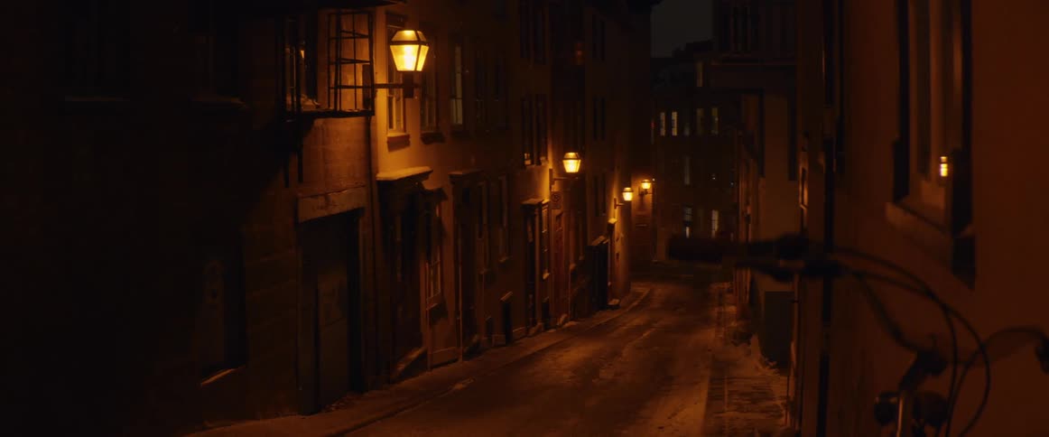 Snow gently falls on a quiet, empty European alleyway illuminated by the warm glow of vintage street lamps at night