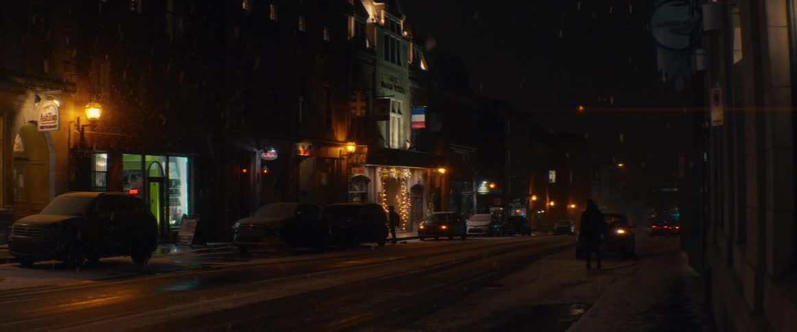 Gentle snow falls over a quiet, warmly lit city street at night as pedestrians walk along the sidewalk