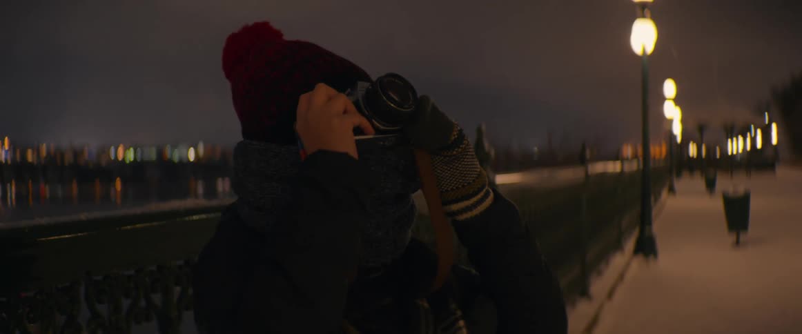 A person bundled in winter clothes takes a picture with a vintage film camera on a snowy boardwalk at night