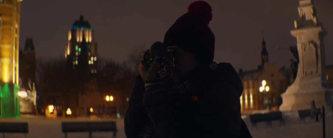 A photographer wearing a beanie and gloves takes a picture of a snowy city park on a cold winter night