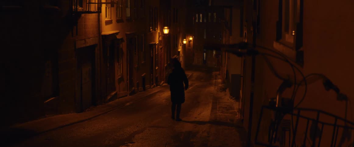 A solitary figure walks away down a narrow, lamp-lit street on a snowy winter night in an old city