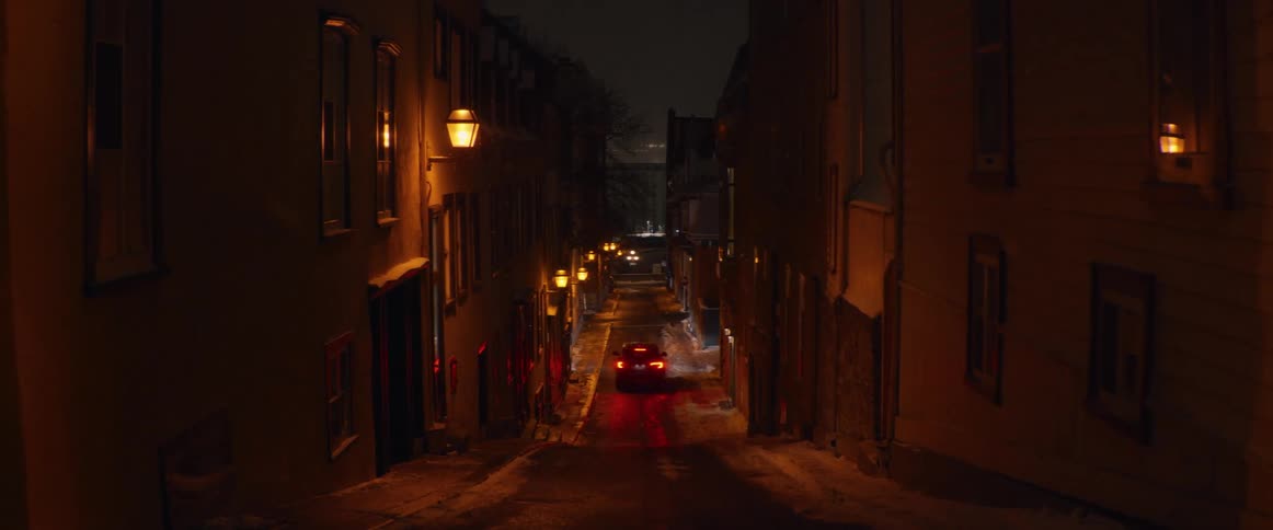 A car's red taillights glow in a narrow, lamp-lit alley on a snowy winter night