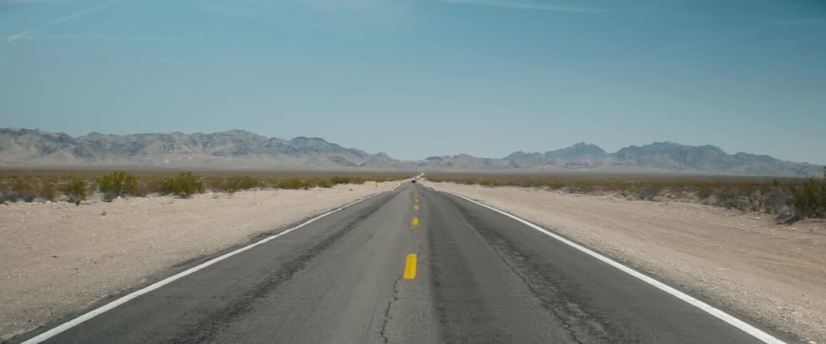 A long, straight desert highway stretches towards distant mountains under a vast, clear sky