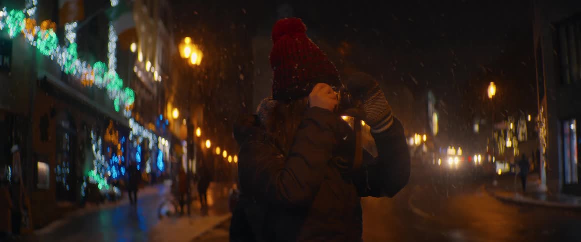 A person in a winter hat and coat photographs the festive holiday lights on a city street during a gentle snowfall at night