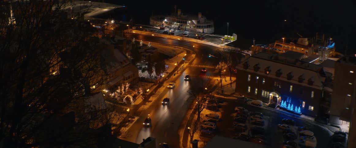 A high-angle view of a charming, snow-covered city at night with festive holiday lights as cars drive along the waterfront