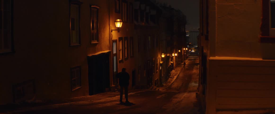 A mysterious person walks alone down a snowy, lamp-lit street in a historic city during a winter night