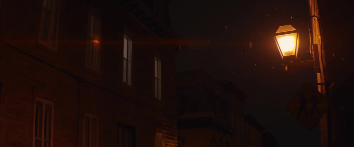 Softly falling snow is illuminated by a vintage street lamp on a quiet city street at night