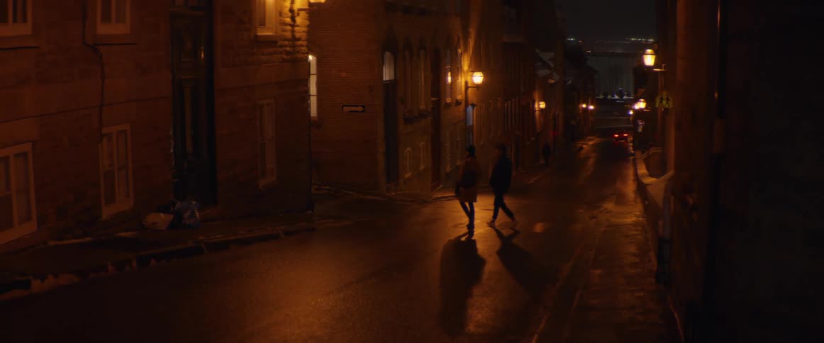 A couple walks in slow motion down a wet, historic city street illuminated by warm lampposts at night