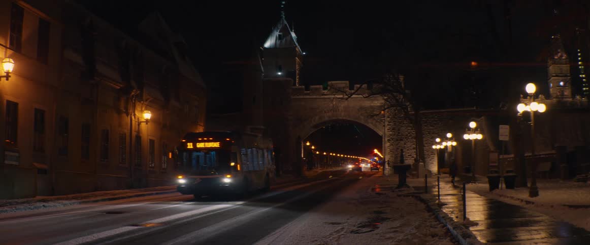 A city bus drives down a snowy street at night, passing by a historic stone gate in Old Quebec