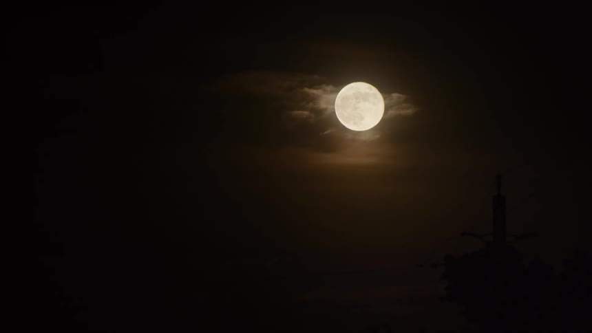 A bright full moon shines through wispy clouds in the dark night sky with the silhouette of a power pole in the foreground