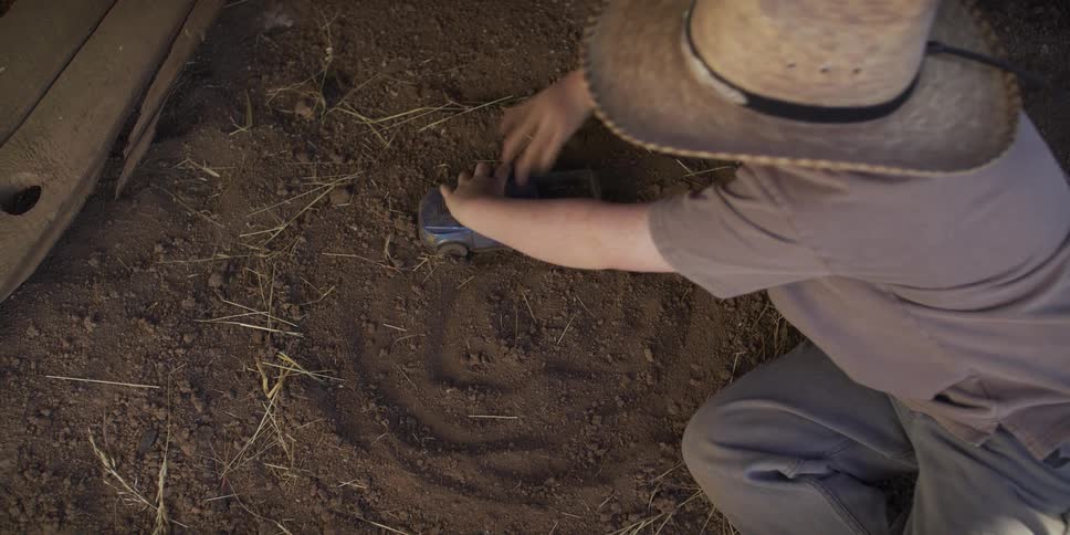 From an overhead view, a young boy in a straw hat creates a spiral track in the dirt with his blue toy truck