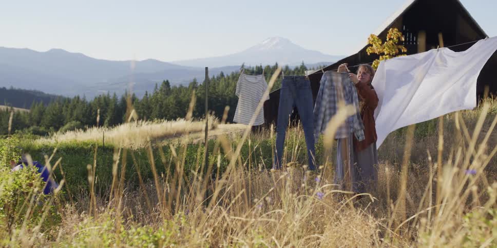 A mother hangs clothes on a clothesline in a rustic field while her son plays, with a large barn and mountain in the distance