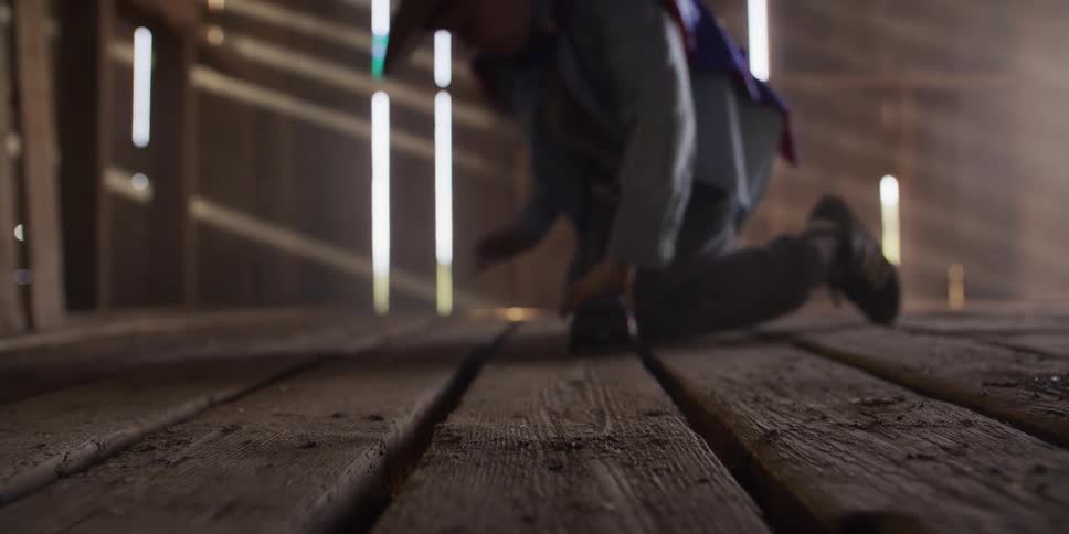 A young boy in a cowboy hat and superhero cape plays with his toy truck in slow motion on the dusty wooden floor of an old barn