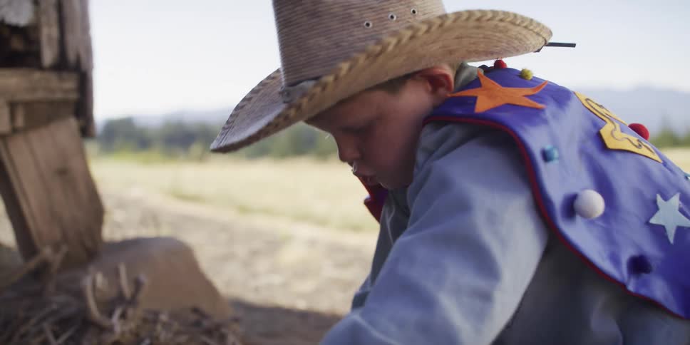 A young boy in a cowboy hat and superhero vest kneels in the dirt and plays with his blue toy truck
