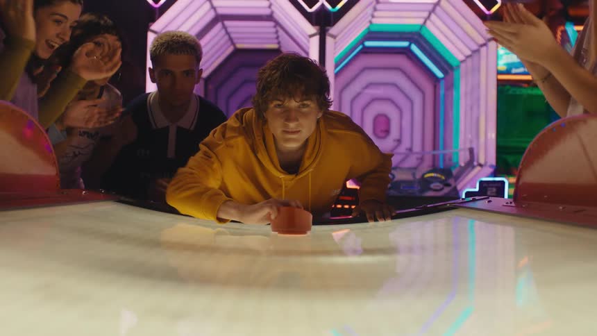 From the perspective of an air hockey table, a focused young man hits the puck towards the camera in slow motion as his friends cheer him on
