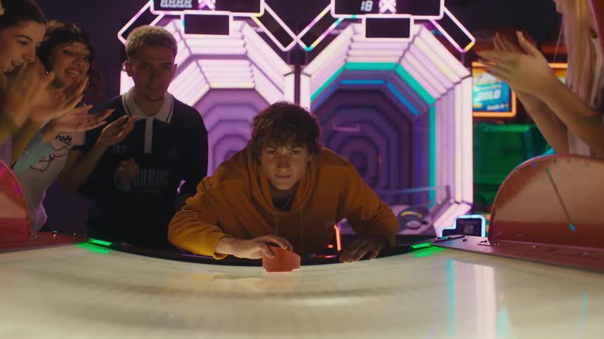 A focused young man hits the air hockey puck in slow motion as his supportive friends cheer him on at a vibrant arcade