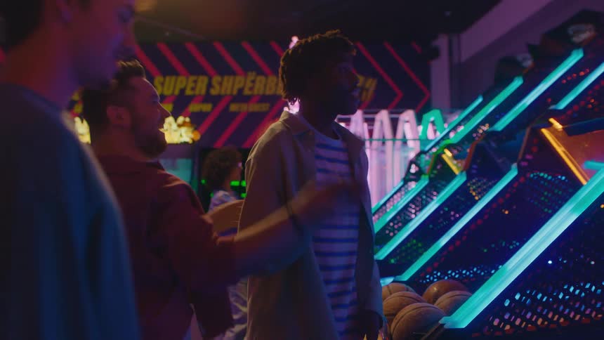 A diverse group of friends laugh and compete while playing a basketball arcade game in a neon-lit game room