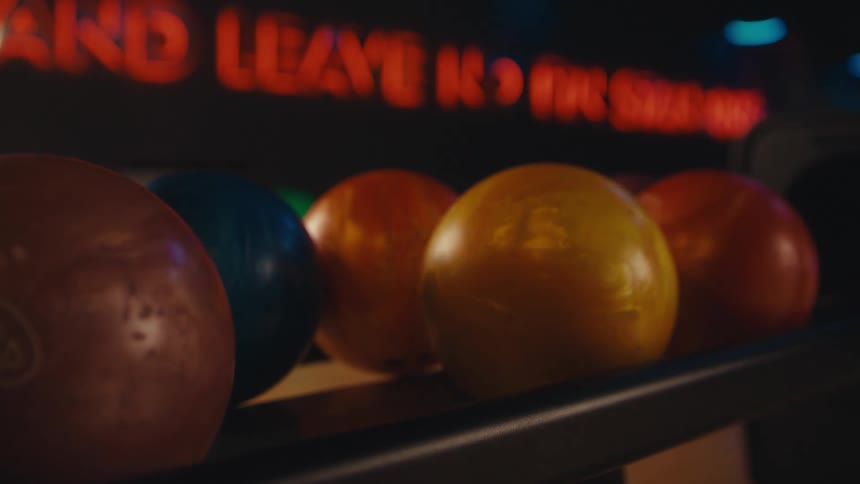 In slow motion, a hand chooses a green bowling ball from the rack at a dimly lit bowling alley