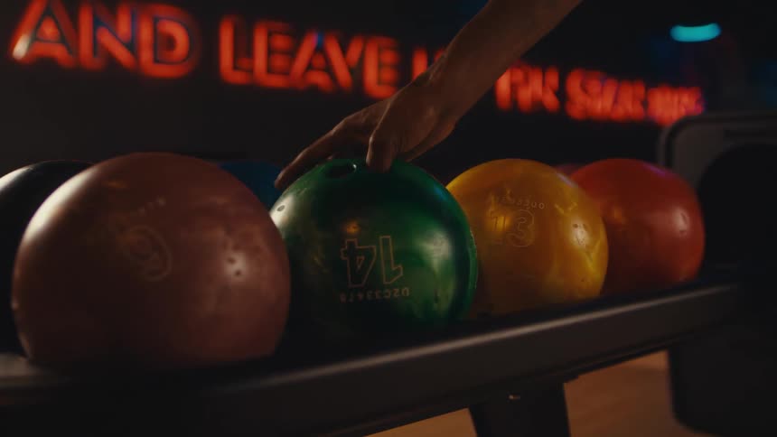 In a retro-style bowling alley, a person's hand selects and picks up a green bowling ball from a rack in slow motion