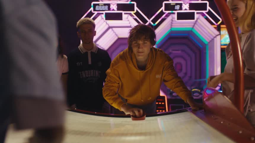 Focused young man in a yellow hoodie competes in a fast-paced game of air hockey as his friends watch and cheer in a vibrant arcade