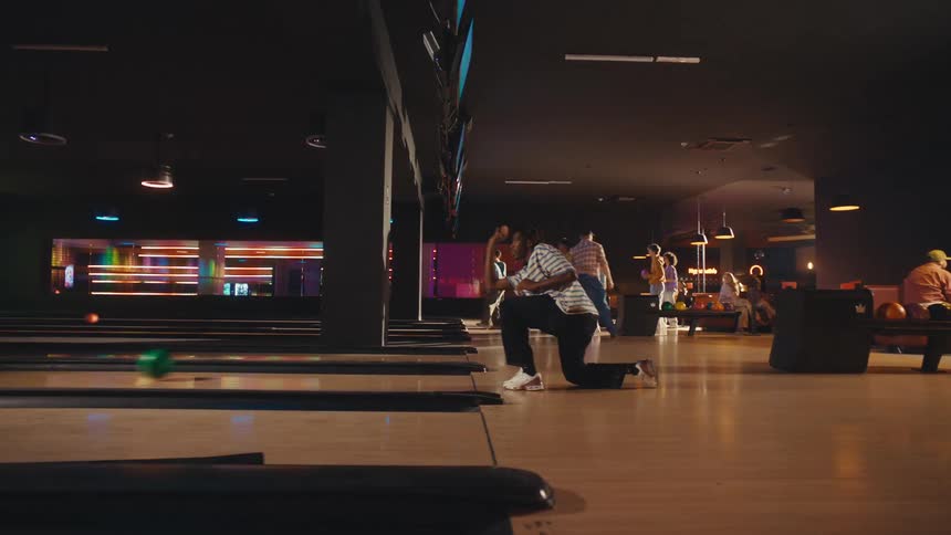 A young Black man skillfully bowls in slow motion, releasing the ball down the lane at a neon-lit alley