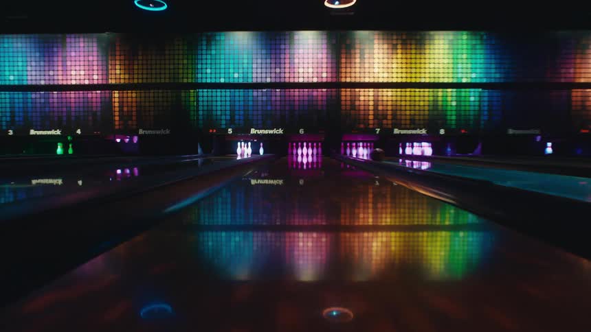 A smooth dolly-in shot down a reflective bowling lane in a dark alley with vibrant, colorful LED lights
