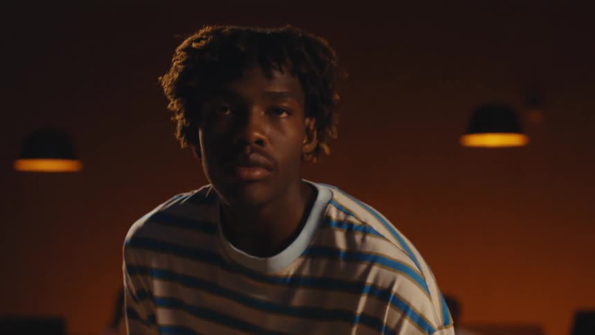 A focused young African American man lowers a green bowling ball from his face and looks intensely at the camera in a bowling alley