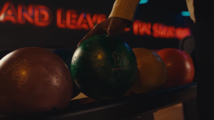 A hand slowly chooses a green bowling ball from a rack in a stylish, dimly lit bowling alley with neon signs