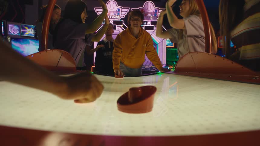 Friends cheer and celebrate in slow motion as a point is scored during an exciting game of air hockey at a neon-lit arcade