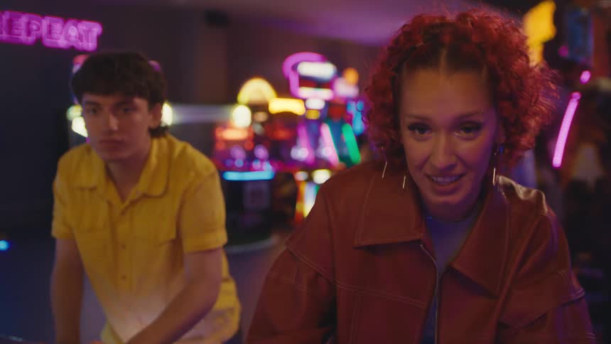 A young woman with red hair and her friend play an arcade game together, smiling competitively in slow motion
