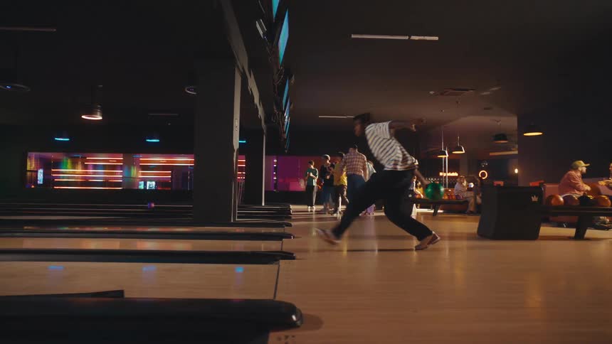 Stylish African American man throws a bowling ball in slow motion as his friends watch and cheer in a neon-lit bowling alley