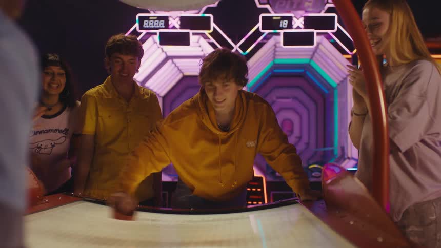 Teenage friends cheer on their two male friends during an exciting game of air hockey in a vibrant, neon-lit arcade