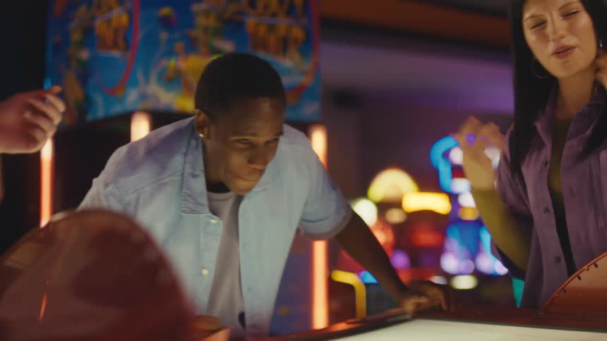An energetic young man laughs and has fun playing a competitive game of air hockey with his friends at an arcade